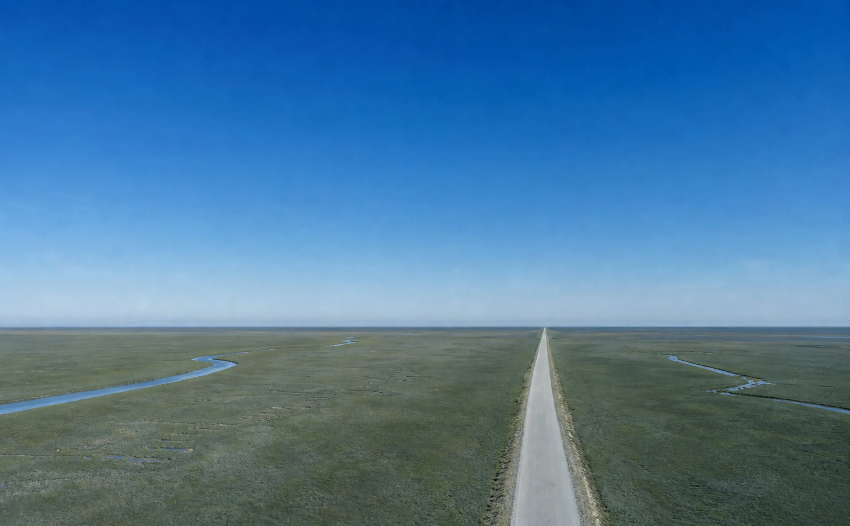 A single narrow road running dead straight across a flat, empty coastal plain under a large clear sky.